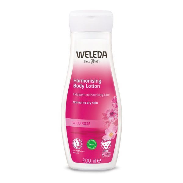  A white, contoured bottle with a pink gradient design, labelled "Weleda Harmonising Body Lotion" with wild rose, for normal to dry skin, in a 200ml volume. The bottle features rose imagery and organic certification logos, set against a plain background.