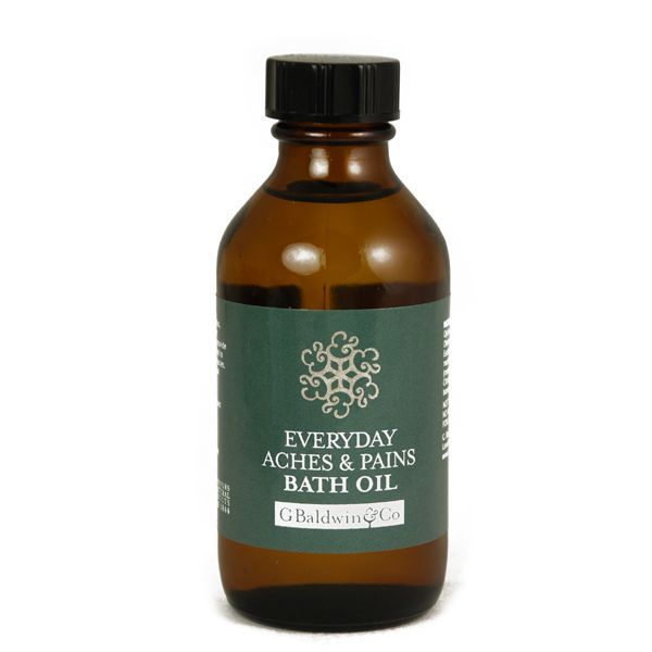  An amber glass bottle with a black cap, labelled "G. Baldwin & Co Everyday Aches & Pains Bath Oil". The label is green with a decorative white emblem, conveying a classic apothecary style, set against a neutral backdrop.